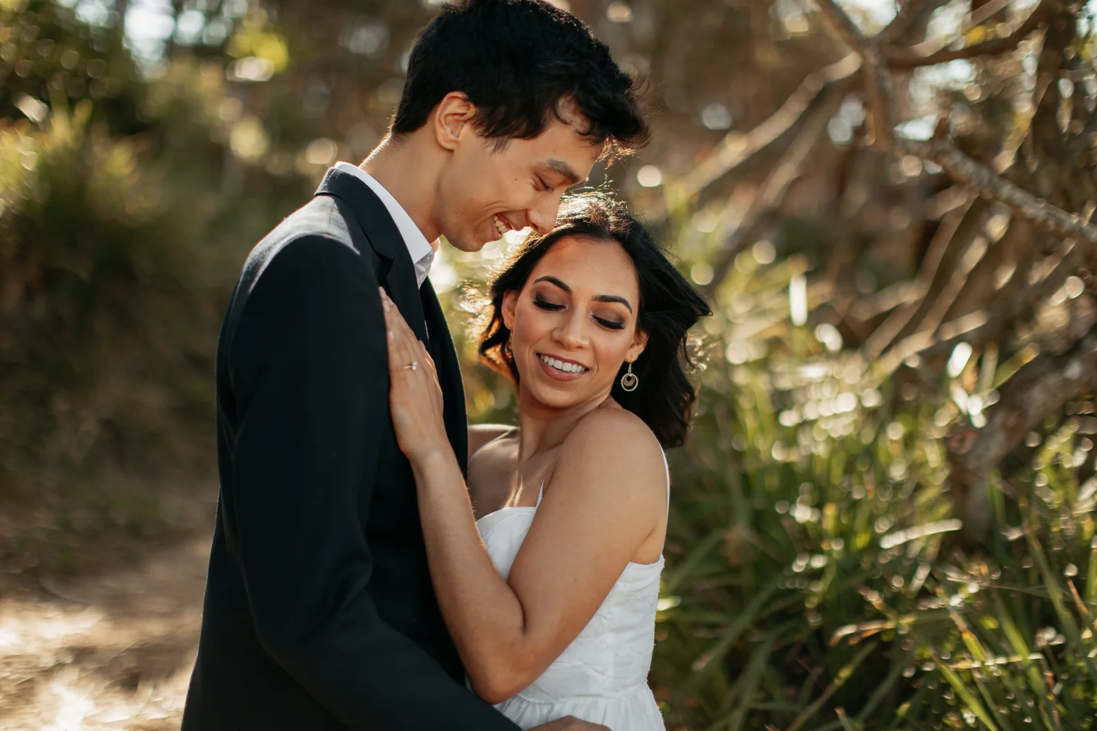 A smiling white-gown bride with her partner beneath the trees