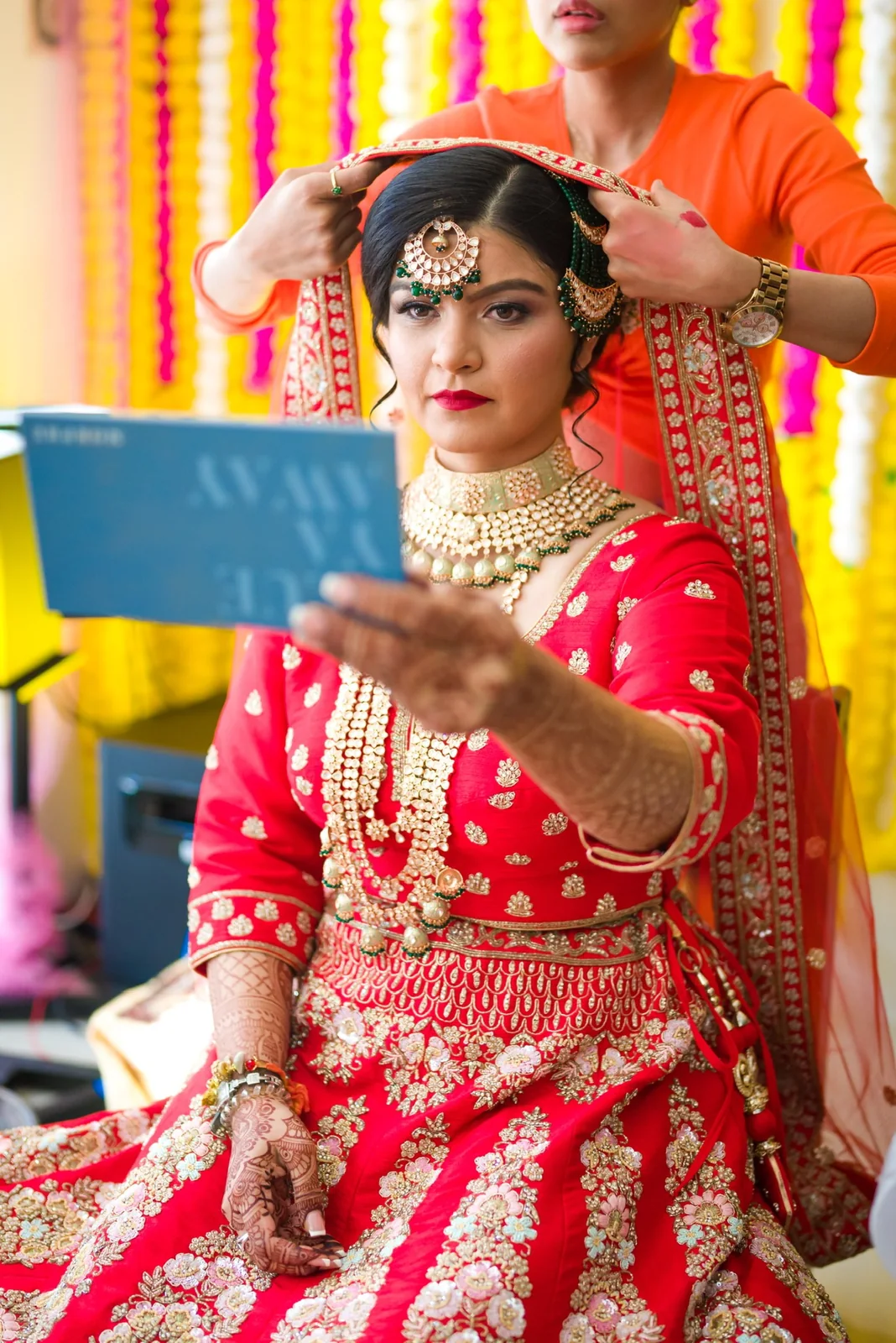 Heera adjusting a red bridal dupatta as the bride checks her hair and makeup in a mirror
