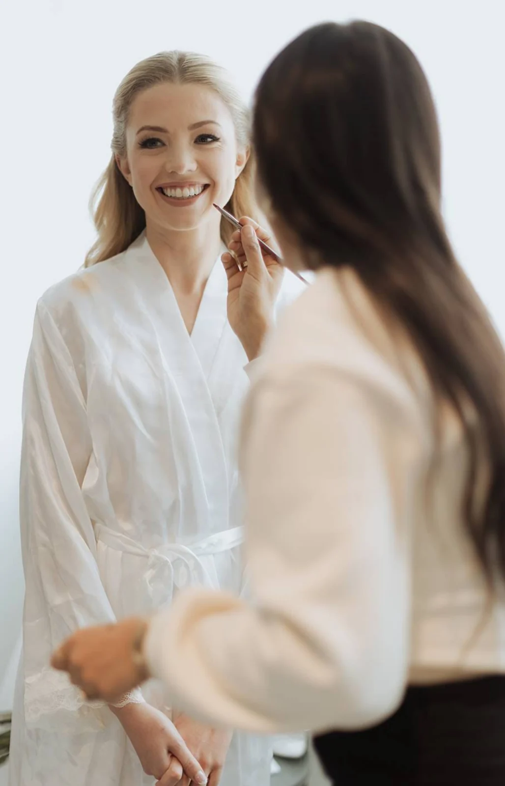 Heera applying finishing makeup to a smiling blonde bride in a white robe