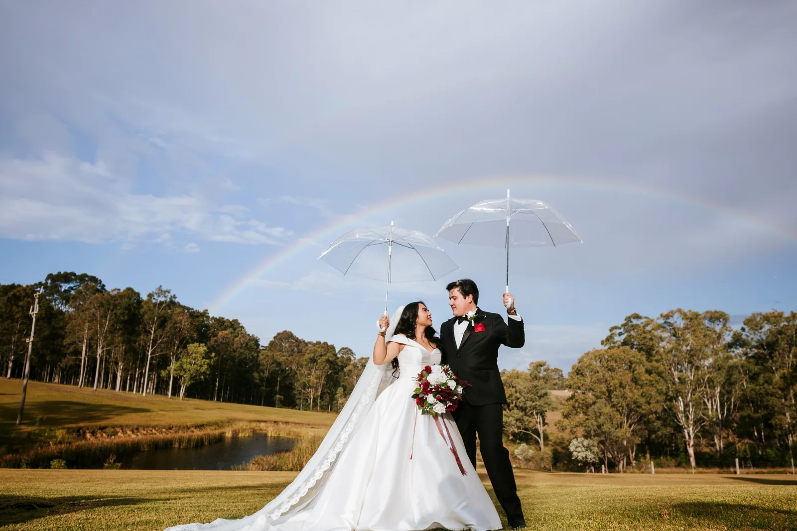 A white-gown couple beneath a clear umbrella on their wedding day