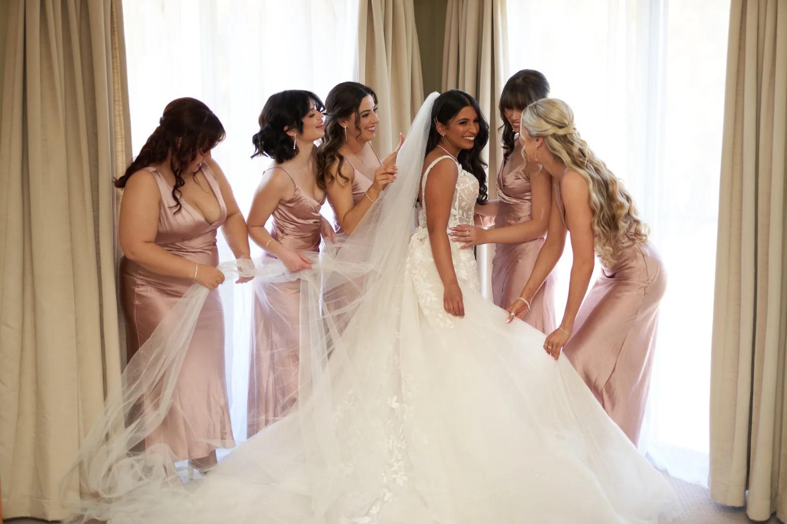 White-gown bride with a veil and softly styled hair, surrounded by bridesmaids in blush gowns