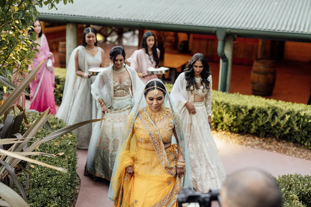 Bride in yellow traditional styling with a veil and jewellery, walking with her bridal party